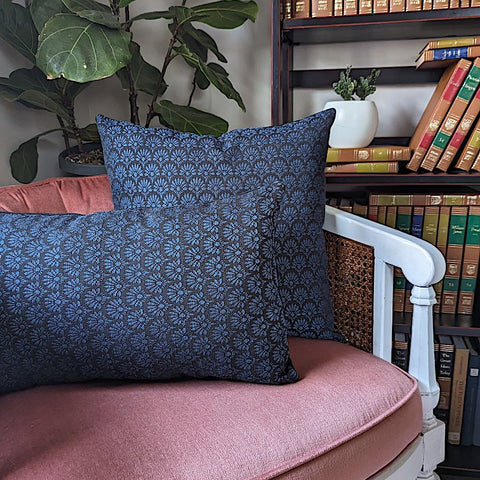two dark blue french pillows with geometric floral designs in reading nook chair with bookshelf and plant in background