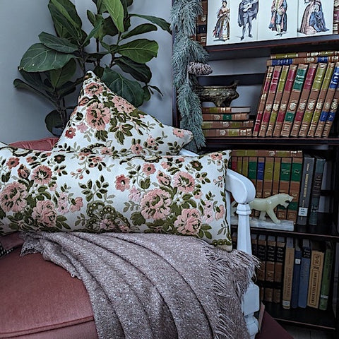 Two french floral pillow cushions on reading nook chair with bookshelf and plant in background
